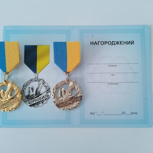 Könnte beinhalten: Drei goldene Medaillen mit blauen und gelben Bändern. Die Medaillen zeigen ein Design eines Atomkraftwerks und das Wort "Chornobyl" in kyrillischer Schrift. Die Medaillen werden auf einem blauen Hintergrund mit dem Text "НАГОРОДЖЕННИЙ" in kyrillischer Schrift angezeigt.