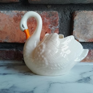 Puede incluir: Figura de cisne de cerámica blanca con pico naranja y detalles de ojos negros. El cisne tiene un cuello curvado y plumas texturizadas. La figura es probablemente un artículo decorativo o una maceta, sobre un muro de ladrillos.