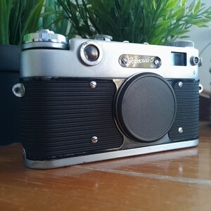 K&ouml;nnte beinhalten: Eine Vintage Zorki 5 Kamera in Silber und Schwarz mit Objektivdeckel. Die Kamera hat einen strukturierten schwarzen Griff und silberne Akzente. Sie steht auf einer Holzoberfl&auml;che, mit gr&uuml;nen Pflanzen im Hintergrund.