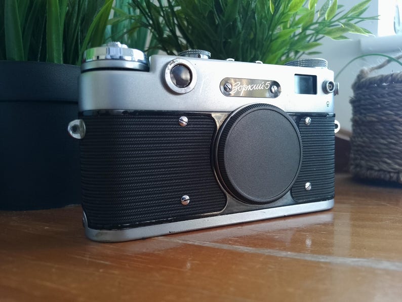 K&ouml;nnte beinhalten: Eine Vintage Zorki 5 Kamera in Silber und Schwarz mit Objektivdeckel. Die Kamera hat einen strukturierten schwarzen Griff und silberne Akzente. Sie steht auf einer Holzoberfl&auml;che, mit gr&uuml;nen Pflanzen im Hintergrund.