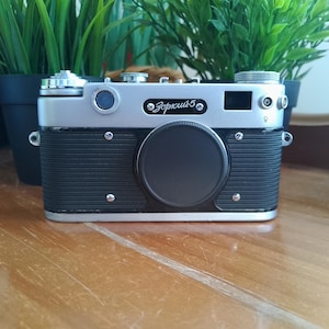 K&ouml;nnte beinhalten: Eine Vintage-Kamera Zorki 5 in Silber und Schwarz mit schwarzem Objektivdeckel. Die Kamera hat eine silberne Oberseite und eine schwarze Vorderseite mit horizontalen Rippen. Die Kamera steht auf einer Holzoberfl&auml;che.
