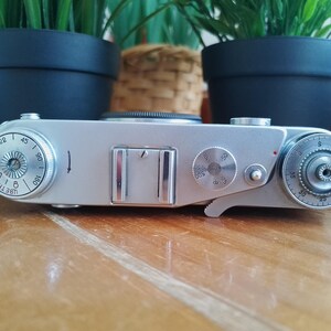 K&ouml;nnte beinhalten: Eine silberne Vintage-Kamera mit mehreren Zifferbl&auml;ttern und Einstellungen. Die Kamera hat einen rechteckigen K&ouml;rper mit einem Objektiv oben und verschiedenen Bedienkn&ouml;pfen. Die Kamera befindet sich auf einer Holzoberfl&auml;che.