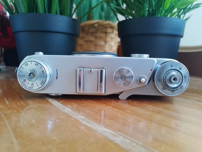 K&ouml;nnte beinhalten: Eine silberne Vintage-Kamera mit mehreren Zifferbl&auml;ttern und Einstellungen. Die Kamera hat einen rechteckigen K&ouml;rper mit einem Objektiv oben und verschiedenen Bedienkn&ouml;pfen. Die Kamera befindet sich auf einer Holzoberfl&auml;che.