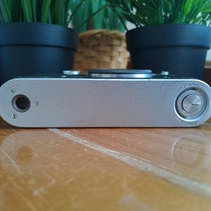 K&ouml;nnte beinhalten: Silbernes Vintage-Kamerageh&auml;use mit schwarzem Objektiv und rundem Zifferblatt. Die Kamera hat eine metallische Oberfl&auml;che und liegt auf einer Holzoberfl&auml;che. Die Kamera ist ein klassisches Design mit Fokus auf Funktionalit&auml;t und Haltbarkeit.