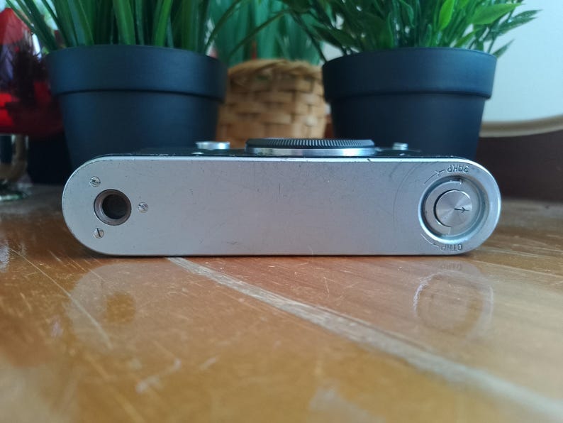 K&ouml;nnte beinhalten: Silbernes Vintage-Kamerageh&auml;use mit schwarzem Objektiv und rundem Zifferblatt. Die Kamera hat eine metallische Oberfl&auml;che und liegt auf einer Holzoberfl&auml;che. Die Kamera ist ein klassisches Design mit Fokus auf Funktionalit&auml;t und Haltbarkeit.