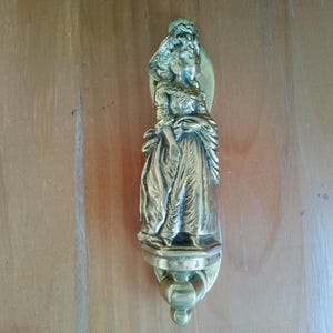 Op de afbeelding: Een messing deurklopper met een gedetailleerde sculptuur van een vrouw in historische kleding. De klopper is gemonteerd op een houten deur en heeft een decoratieve basis met een gebogen handvat.