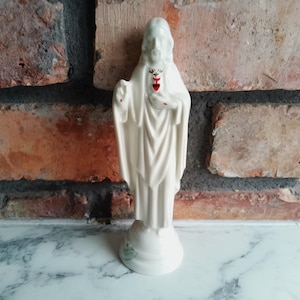 Puede incluir: Estatua de cerámica blanca de Jesucristo, de pie sobre una base redonda. La estatua tiene un rostro detallado y túnicas fluidas. Un corazón rojo es visible en el pecho. El fondo es una pared de ladrillos.