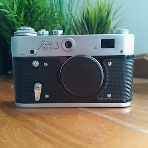 May include: A silver and black FED 3 rangefinder camera featuring a textured black body, a central circular lens cap, a silver top plate with dials, and the text FED 3 in Cyrillic and Latin characters printed on the front of the camera body.