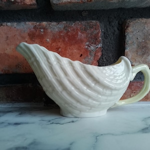 Pode incluir: Molheira de cerâmica creme com um desenho de concha. A molheira tem uma pega e um bico, e assenta numa superfície de mármore. O fundo é uma parede de tijolo. Esta molheira é um artigo de cozinha.