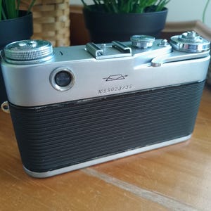 K&ouml;nnte beinhalten: Eine Vintage-Kamera in Silber und Schwarz mit einem strukturierten schwarzen Griff. Die Kamera hat mehrere silberne Drehkn&ouml;pfe und ein Objektiv auf der Vorderseite. Die Seriennummer "№ 59023/35" ist auf der Vorderseite aufgedruckt. Ein klassisches Beispiel f&uuml;r Fotoausr&uuml;stung.