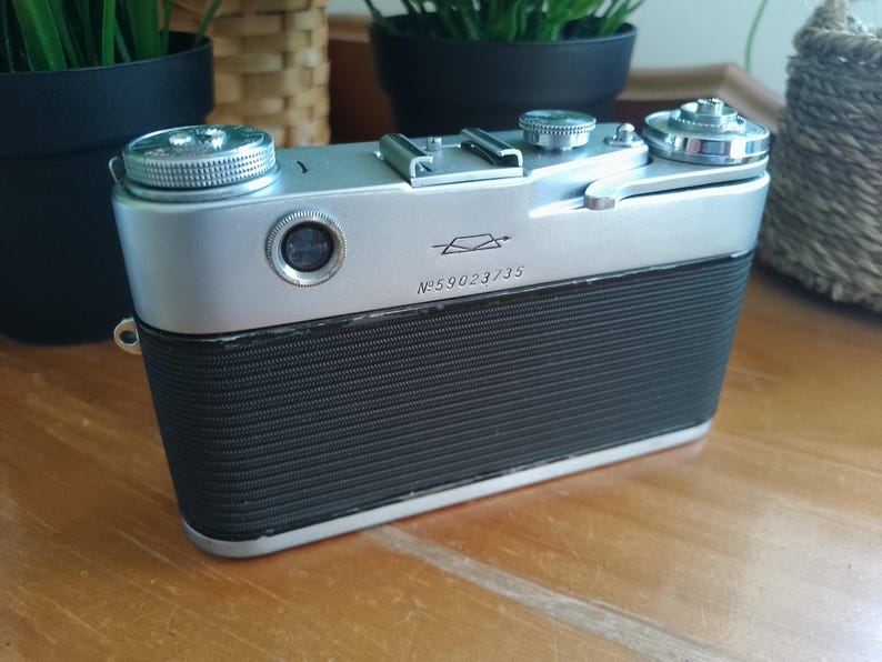K&ouml;nnte beinhalten: Eine Vintage-Kamera in Silber und Schwarz mit einem strukturierten schwarzen Griff. Die Kamera hat mehrere silberne Drehkn&ouml;pfe und ein Objektiv auf der Vorderseite. Die Seriennummer "№ 59023/35" ist auf der Vorderseite aufgedruckt. Ein klassisches Beispiel f&uuml;r Fotoausr&uuml;stung.