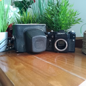 May include: A black Zenit camera and its leather case are on a wooden surface. The camera has a silver lens ring and the word "ZENIT" is printed on the top. The case is black and has a square shape.