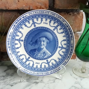 May include: A decorative plate with a blue and white design. The plate features a portrait of Queen Elizabeth the Queen Mother, with text that reads "To Celebrate the Life of Queen Elizabeth the Queen Mother 1900-2002". The plate is displayed on a clear stand.