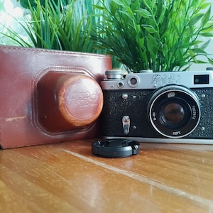 May include: A vintage black and silver FED 2 camera with a lens cap, next to a brown leather camera case. The camera has a prominent lens and several dials. Green plants are in the background.