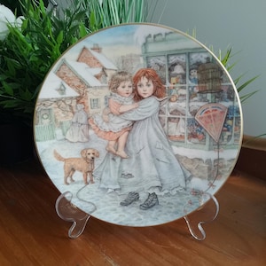 Può includere: Un piatto decorativo rotondo in porcellana con bordo dorato, caratterizzato da un'illustrazione dettagliata di un bambino con un lungo abito grigio che tiene in braccio un bambino più piccolo con un abito color pesca, con un cagnolino beige.