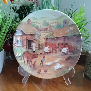 Pode incluir: Prato decorativo com uma cena de fazenda pastoral com vacas, galinhas e pessoas. O prato tem uma ilustração detalhada de uma vila rural com uma casa com telhado de colmo e uma igreja. O prato é exibido em um suporte transparente.