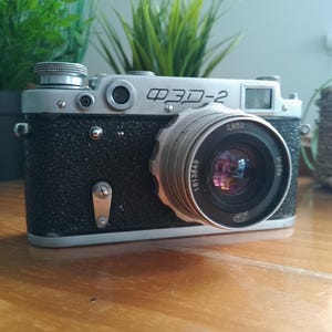 May include: A vintage FED-2 rangefinder camera with a black textured body and silver metal top plate. The lens is marked with the text W-20M, 1613849, and 2,8/52. The camera features a silver shutter release button, a viewfinder, and a small lever.