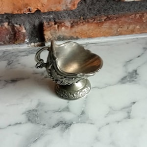 Puede incluir: Una pequeña salsera plateada y ornamentada, con asa y base decorativa. El recipiente tiene un borde festoneado y está sobre una superficie de mármol. El fondo incluye una pared de ladrillo.