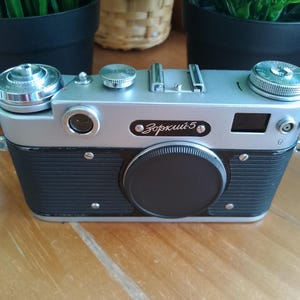 K&ouml;nnte beinhalten: Eine Vintage-Kamera Zorki 5 in Silber und Schwarz mit schwarzem Objektivdeckel. Die Kamera hat eine silberne Oberseite und einen schwarzen, strukturierten K&ouml;rper. Die Kamera hat mehrere Drehkn&ouml;pfe und Tasten. Die Kamera steht auf einer Holzoberfl&auml;che.
