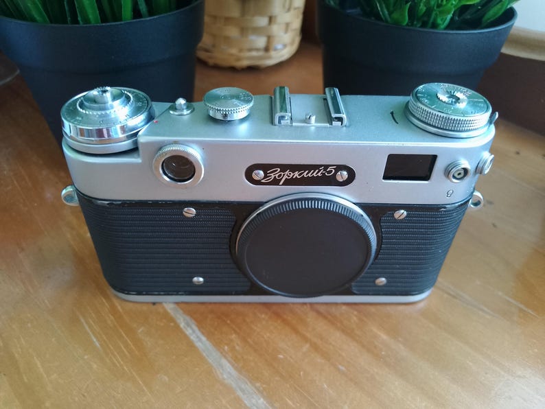 K&ouml;nnte beinhalten: Eine Vintage-Kamera Zorki 5 in Silber und Schwarz mit schwarzem Objektivdeckel. Die Kamera hat eine silberne Oberseite und einen schwarzen, strukturierten K&ouml;rper. Die Kamera hat mehrere Drehkn&ouml;pfe und Tasten. Die Kamera steht auf einer Holzoberfl&auml;che.