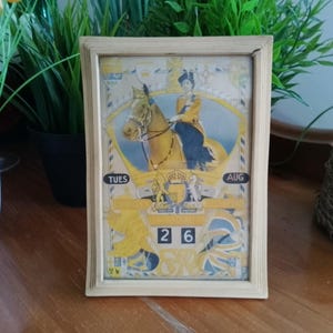 May include: A framed vintage calendar print featuring a woman on horseback in yellow and blue attire. The calendar displays the date August 26th, with the days of the week and month in yellow and blue. The frame is a light wood color.