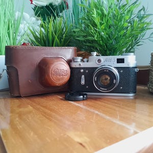 May include: A vintage black and silver camera with a lens cap, next to a brown leather camera case. The camera has the text "Зоркий 3" on the top. The camera and case are on a wooden surface, with green plants in the background.