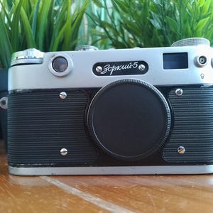 K&ouml;nnte beinhalten: Eine Vintage Zorki-5 Kamera mit silbernem Oberteil und schwarzem Geh&auml;use. Das Objektiv ist mit einer schwarzen Kappe abgedeckt. Die Kamera hat den Text "Зоркий-5" auf der Vorderseite. Die Kamera steht auf einer Holzoberfl&auml;che.