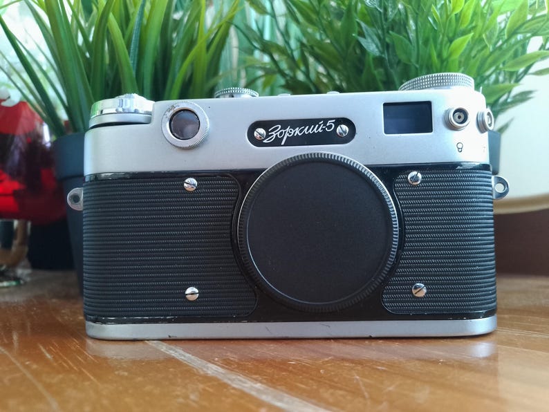 K&ouml;nnte beinhalten: Eine Vintage Zorki-5 Kamera mit silbernem Oberteil und schwarzem Geh&auml;use. Das Objektiv ist mit einer schwarzen Kappe abgedeckt. Die Kamera hat den Text "Зоркий-5" auf der Vorderseite. Die Kamera steht auf einer Holzoberfl&auml;che.