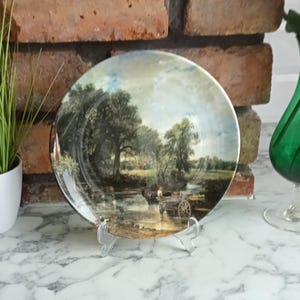 May include: A decorative plate featuring a landscape painting with trees, a river, and a horse-drawn cart. The plate is displayed on a clear plastic stand, with a brick wall and a green vase in the background.