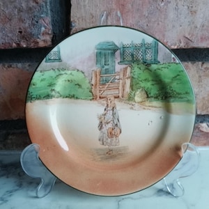 Può includere: Un piatto decorativo con una scena pastorale. Il piatto presenta una figura in un vestito, un cancello e una casa. Il piatto ha un centro color crema con un bordo arancione e un bordo verde.