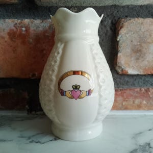 May include: A white ceramic vase with a scalloped rim and a textured pattern. The vase features a gold and pink Claddagh design with a heart, crown, and hands. The vase is approximately 15 cm tall.