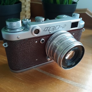 Może przedstawiać: Aparat dalmierzowy vintage FED-2 ze srebrną metalową płytką górną i korpusem z teksturowanej brązowej skóry. Aparat posiada srebrny obiektyw z widocznymi oznaczeniami przysłony i ostrości oraz pokrętło przewijania filmu na górnej części.