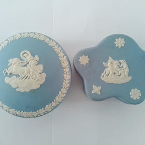 May include: Two blue and white ceramic trinket boxes. One box is round with a white design of a chariot pulled by two horses. The other box is shaped like a flower with a white design of a woman riding a horse.