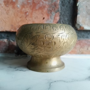 Może przedstawiać: Mała, złota miedziana miska z misternymi wzorami kwiatowymi wygrawerowanymi na powierzchni. Miska ma szeroką, zaokrągloną podstawę i rozszerzający się brzeg.