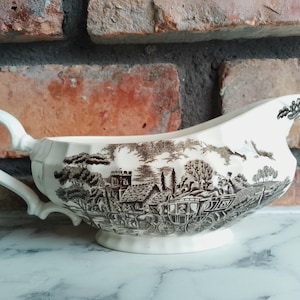 Może przedstawiać: Kremowa łódka do sosu z szczegółowym czarno-białym wzorem transferowym przedstawiającym wiejską scenę z powozem konnym. Łódka do sosu ma zakrzywiony uchwyt i dekoracyjny dzióbek.