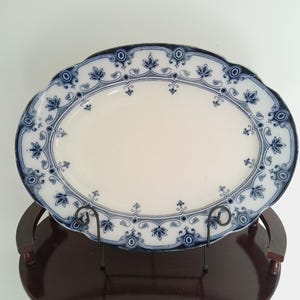 Pode incluir: Travessa oval para servir com centro branco e borda floral azul. A borda tem uma borda ondulada. A travessa é exibida em um suporte marrom escuro. Uma peça clássica para servir comida.