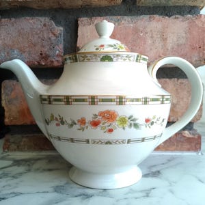 May include: A white ceramic teapot with a lid, decorated with floral and geometric patterns in orange, green, and yellow. The teapot has a curved handle and spout, and sits on a small pedestal base. The background includes a brick wall.