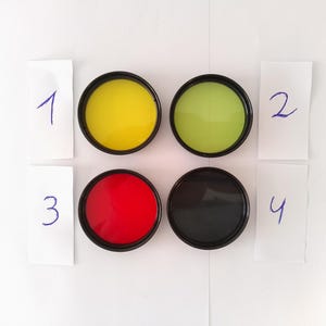Op de afbeelding: Vier ronde containers met deksels, genummerd van 1 tot en met 4. De containers zijn gevuld met gele, groene, rode en zwarte verf.