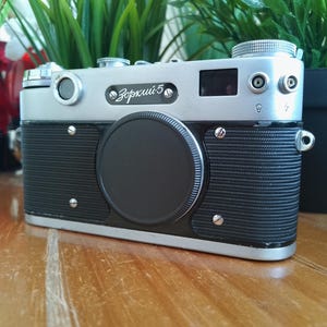 K&ouml;nnte beinhalten: Eine Vintage Zorki 5 Kamera in Silber und Schwarz mit schwarzem Objektivdeckel. Die Kamera hat einen geriffelten schwarzen Griff und eine silberne Oberseite mit Kn&ouml;pfen und R&auml;dern. Der Text "Зоркий 5" ist auf der Vorderseite zu sehen.