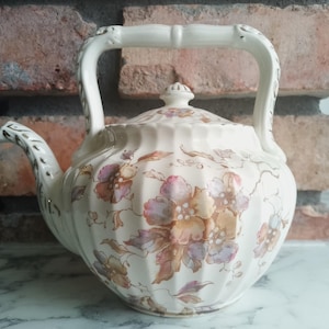 Könnte beinhalten: Cremefarbene Keramik-Teekanne mit einem Blumenmuster in Rosa, Lila und Beige. Die Teekanne hat einen gebogenen Ausguss, einen Griff im Bambus-Stil und einen passenden Deckel. Ein dekorativer Artikel für Teeliebhaber.
