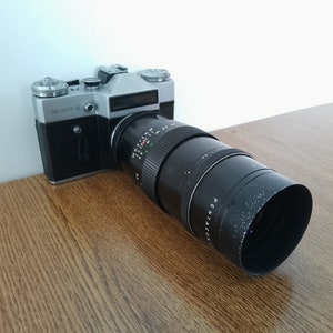 May include: A black and silver Zenit-E film camera with a Pentacon 135mm f/3.5 lens attached. The camera is on a wooden surface.