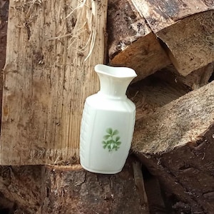 Puede incluir: Un pequeño jarrón de cerámica blanca con la parte superior acampanada y un diseño floral verde. El jarrón tiene un patrón texturizado en los lados. El jarrón mide aproximadamente 15 cm de alto.