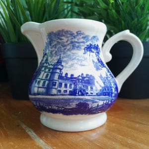 Puede incluir: Una pequeña jarra de cerámica blanca con un diseño de paisaje azul y blanco. La jarra tiene un asa y un pico, y el diseño representa un edificio con árboles y una masa de agua. La jarra está sobre una superficie de madera.
