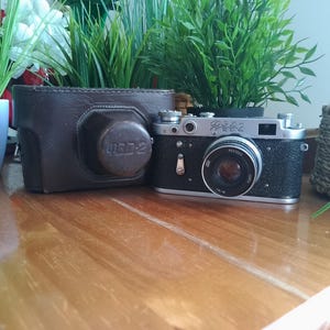 May include: A classic black and silver rangefinder camera with a brown leather case. The camera features a large lens and several dials and buttons. The leather case has a circular lens cover. The camera is positioned on a wooden surface.