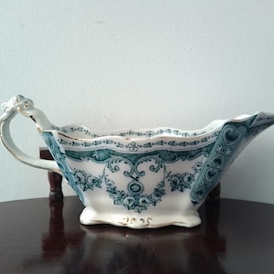 May include: Antique white and teal porcelain gravy boat with a floral design. The boat features a handle and a spout, with gold trim accents. The design includes floral patterns and decorative borders, showcasing a vintage aesthetic.