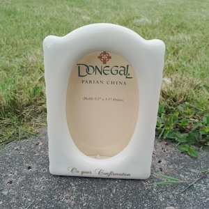 May include: A white ceramic photo frame with a curved top and an oval opening. The frame has the text "Donegal Parian China" and "On your Confirmation". The frame is designed to hold a 3.5" x 5.5" print.