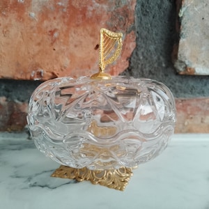 May include: A clear, cut-glass trinket box with a gold-colored harp finial. The box sits on a gold-colored, ornate base. The background features a brick wall and a white surface.