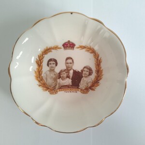 Puede incluir: Un plato de cerámica blanca con un borde dorado y una imagen de la Familia Real de la Coronación de 1937. El plato tiene una corona de hojas alrededor de la imagen y las palabras "The Royal Family Coronation 1937 Souvenir" impresas en él.