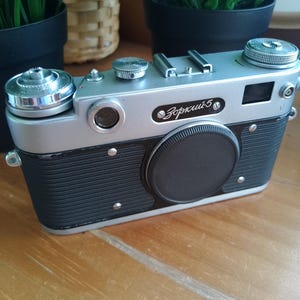 K&ouml;nnte beinhalten: Eine Vintage-Kamera Zorki-5 in Silber und Schwarz mit Objektivdeckel. Die Kamera hat eine silberne Oberseite und einen schwarzen K&ouml;rper mit strukturierter Grifffl&auml;che. Der Name "Зоркий-5" ist auf der Vorderseite aufgedruckt. Ein klassisches Fotoequipment.