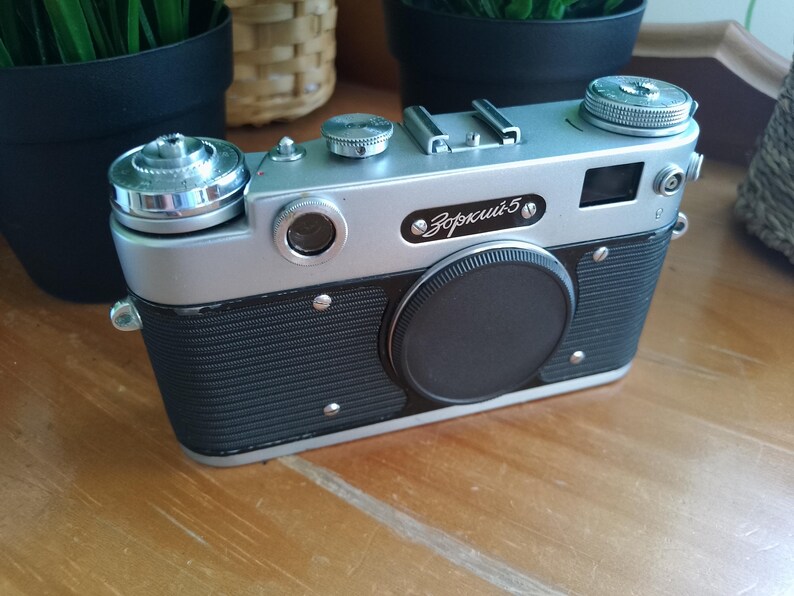 K&ouml;nnte beinhalten: Eine Vintage-Kamera Zorki-5 in Silber und Schwarz mit Objektivdeckel. Die Kamera hat eine silberne Oberseite und einen schwarzen K&ouml;rper mit strukturierter Grifffl&auml;che. Der Name "Зоркий-5" ist auf der Vorderseite aufgedruckt. Ein klassisches Fotoequipment.